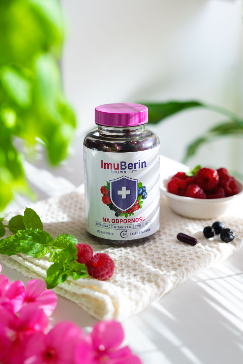 Natural Pharmaceuticals ImuBerin IToidulisand ImuBerin immuunsüsteemi tugevdamiseks esteetilises keskkonnas – vitamiinide C, D, tsingi ja seleeniga kapslite pudel, asetatud heledale rätikule, mis on kaunistatud vaarikate, piparmündi ja lilledega, taustal puuviljade kauss ja rohelised lehed.